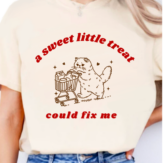 Beige t-shirt with a graphic of a cat with a shopping cart and text 'a sweet little treat could fix me' on a white background.